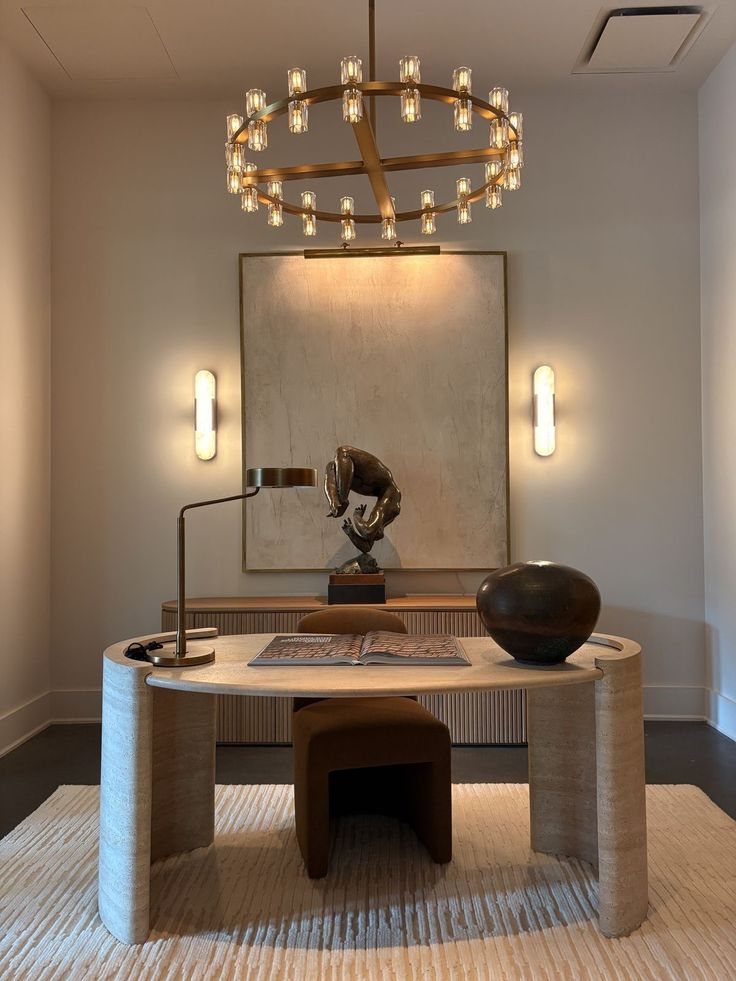 A travertine desk under a brass chandelier, with a bronze sculpture, an open book and a dark ceramic vase.