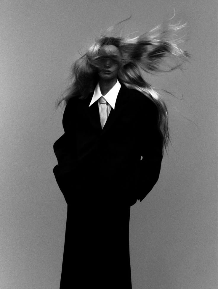 Black and white editorial portrait of a model in a long black coat, her hair blowing across her face.
